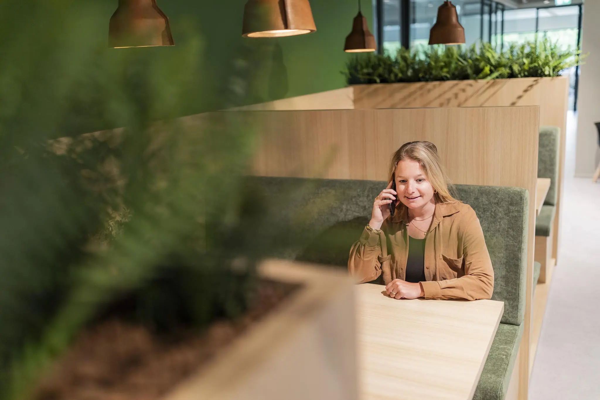 Vrouw voert telefoongesprek in een modern kantoor met groene planten en houten werkplekken.