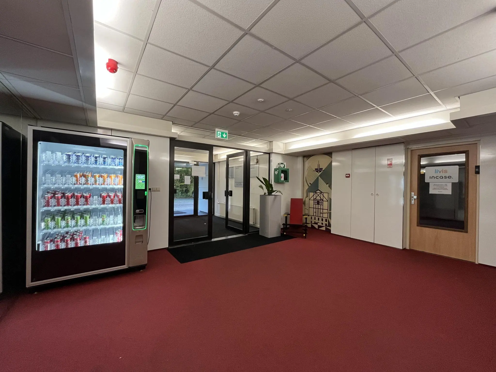 Hal met frisdrankautomaat, glazen deuren, een deur met Livis Incase-logo en rode vloerbedekking in een kantoorgebouw aan de Industrieweg.