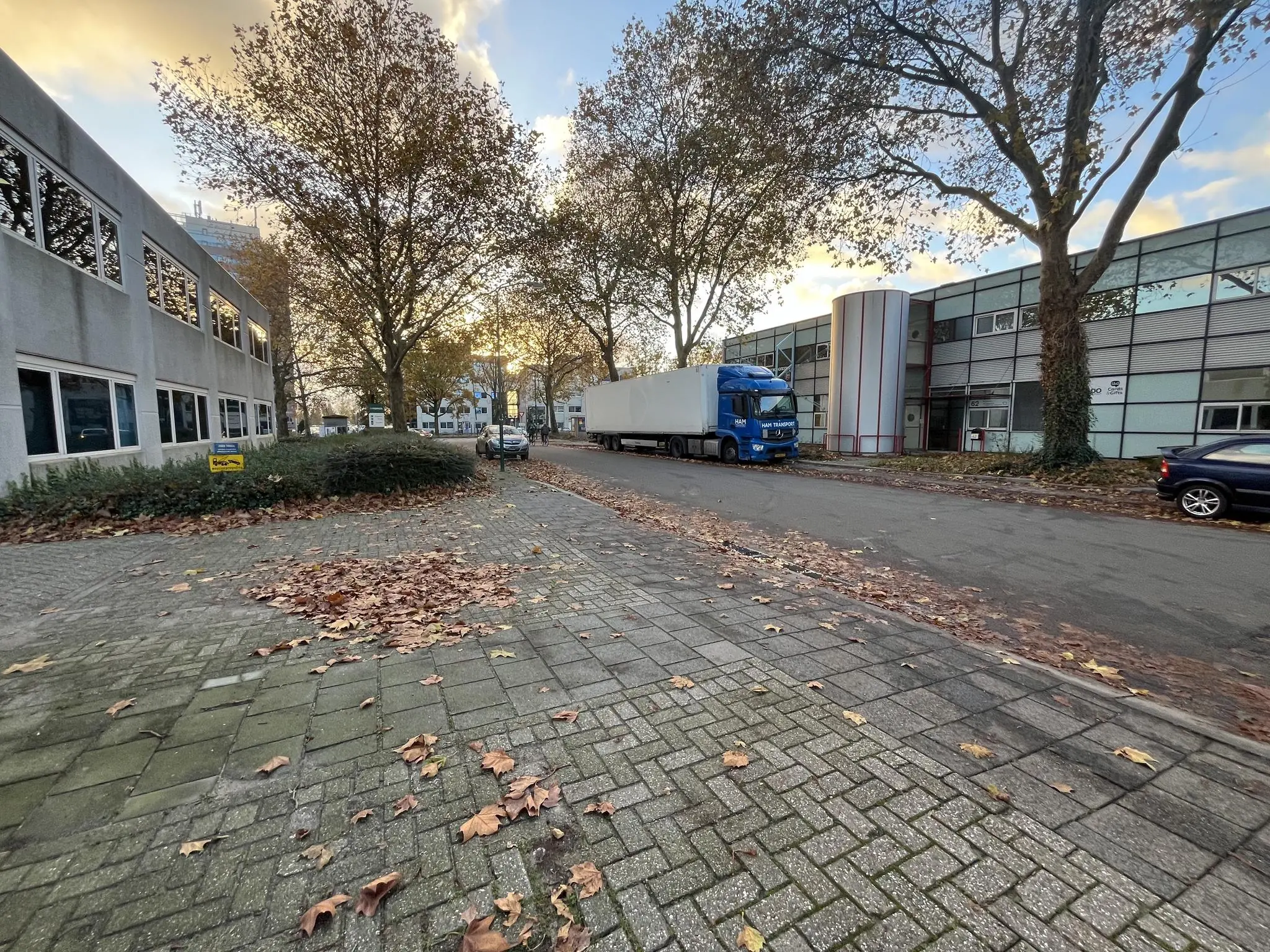 Een industrieweg met kantoorpanden, geparkeerde auto's en een vrachtwagen op een herfstige dag.
