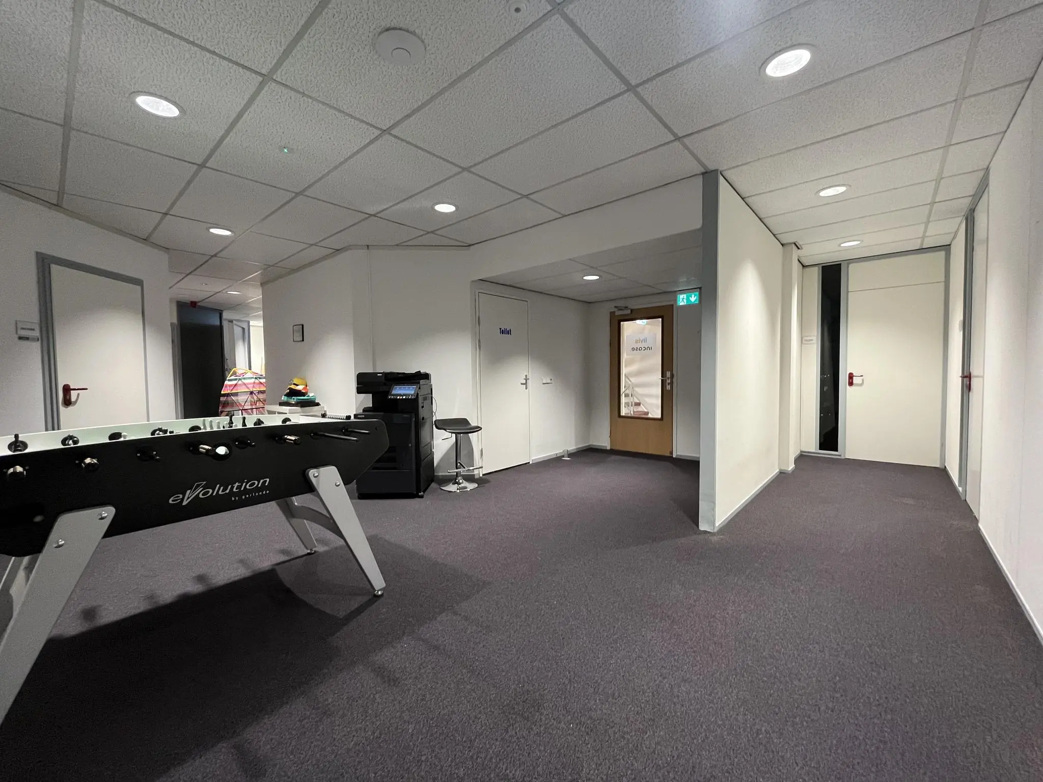 Kantoorhal met een tafelvoetbaltafel, printer en deurlabels waaronder 'Toilet' en 'Inhouse Inside'.