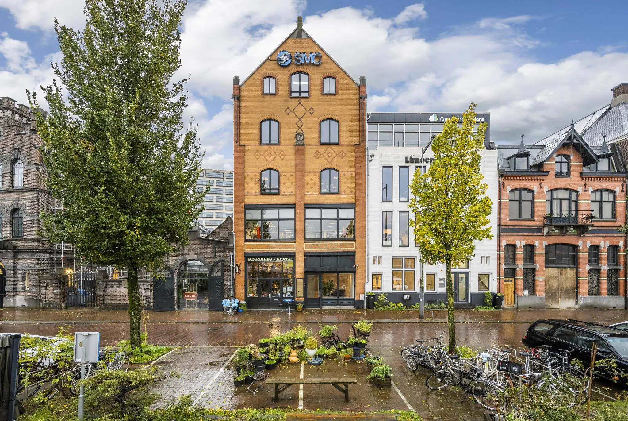 Voorgevel van een karakteristiek pand aan de De Ruijterkade in Amsterdam met bomen, fietsen en een nat wegdek op de voorgrond.