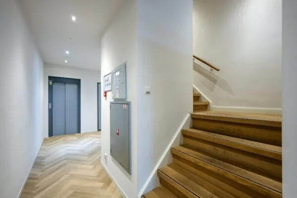 Hal met houten trap, liftdeuren en informatieborden in een modern gebouw aan de De Ruijterkade.