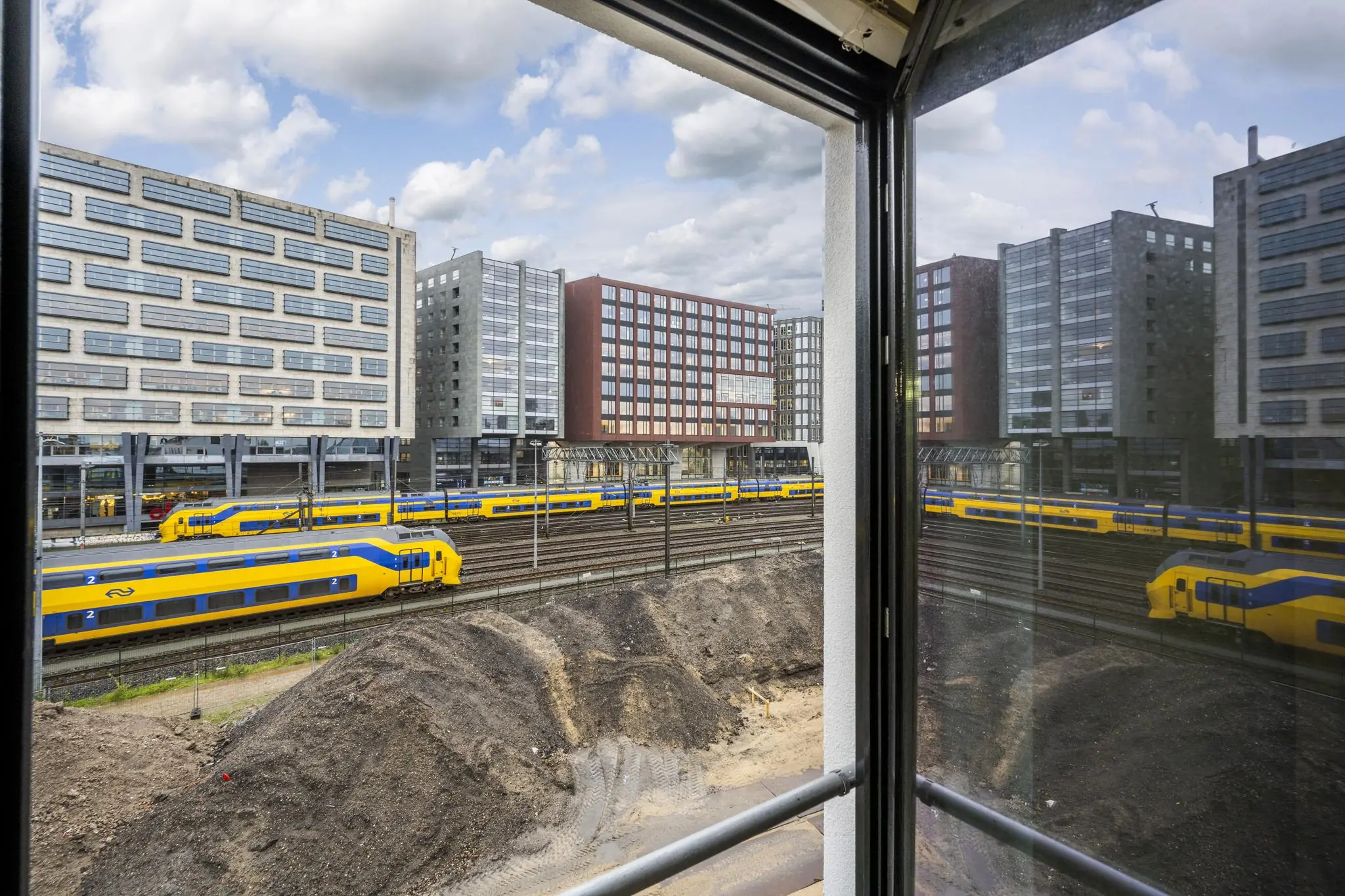 Uitzicht vanaf een raam op De Ruijterkade met spoorlijnen, passerende treinen en omliggende kantoorgebouwen.
