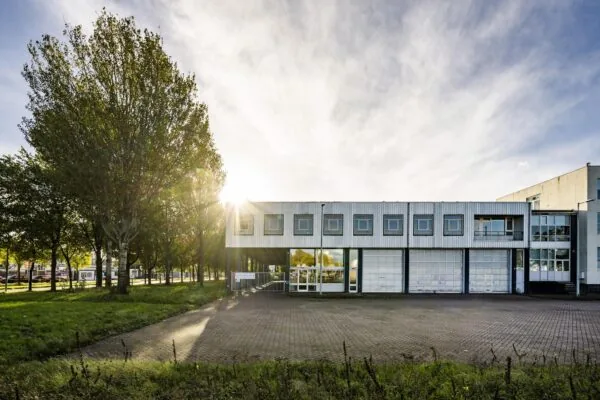 Wit industrieel kantoorgebouw aan de Van der Madeweg met zonlicht tussen de bomen en een lege parkeerplaats op de voorgrond.