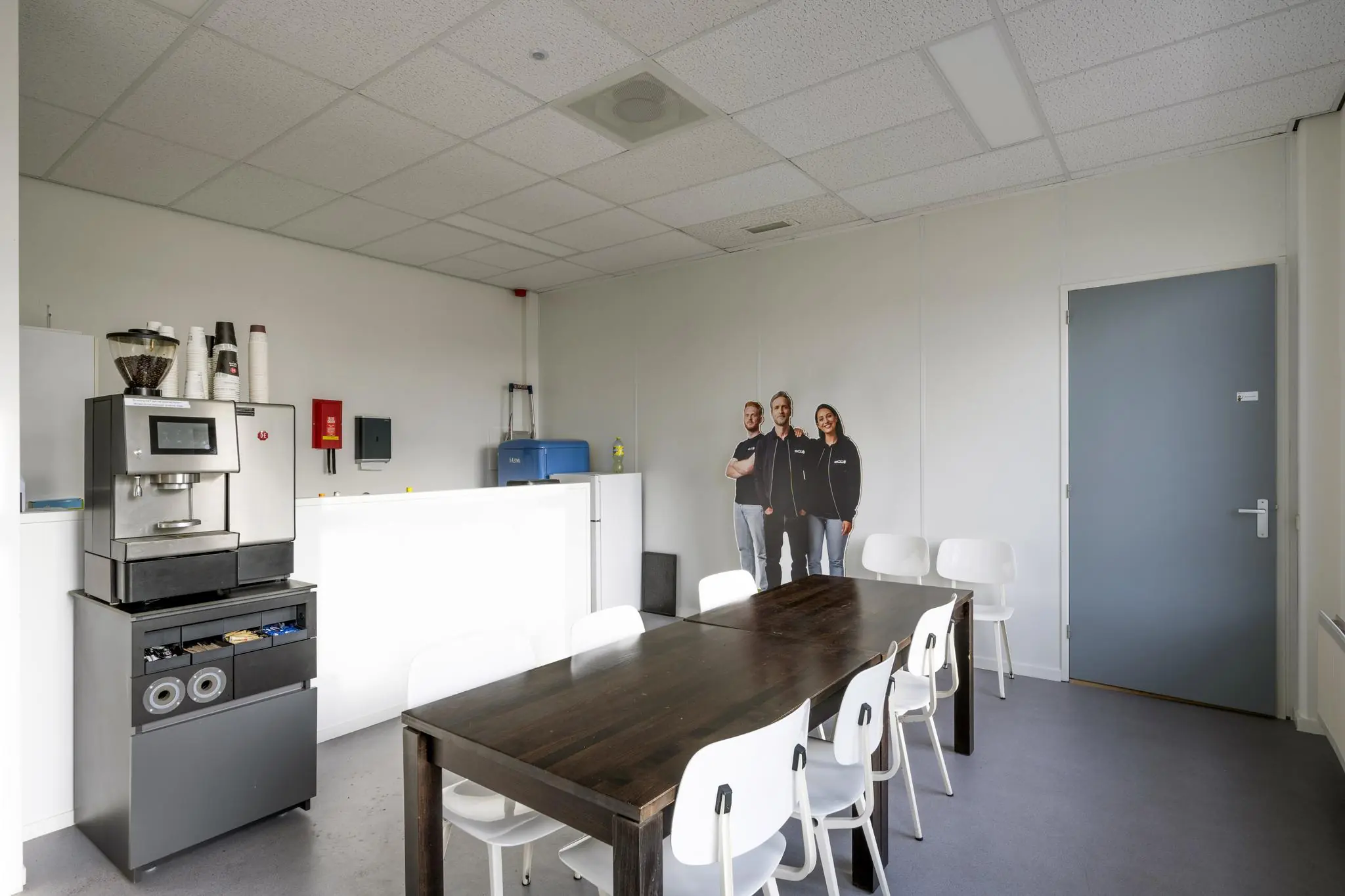 Kantineruimte aan de Van der Madeweg met koffiemachine, houten tafel, witte stoelen en een kartonnen figuur van drie mensen tegen de muur.