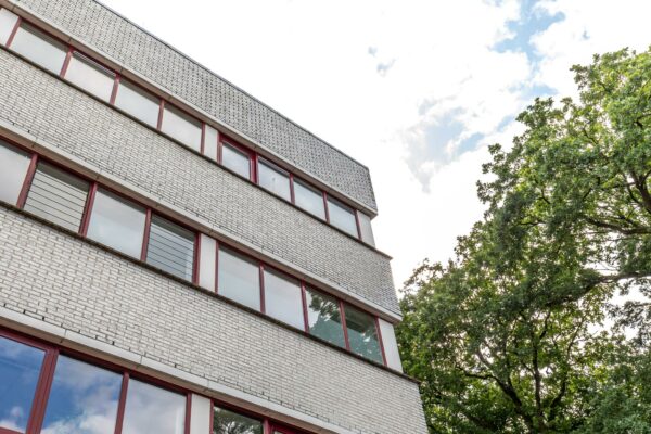 Vooraanzicht van een lichtgrijs kantoorgebouw aan de Birkstraat met rode raamkozijnen en omliggende bomen.