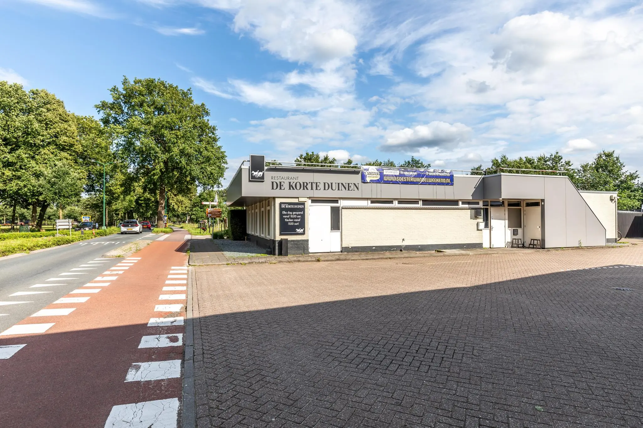 Restaurant De Korte Duinen aan de Birkstraat met naastgelegen fietspad en parkeerplaats op een zonnige dag.