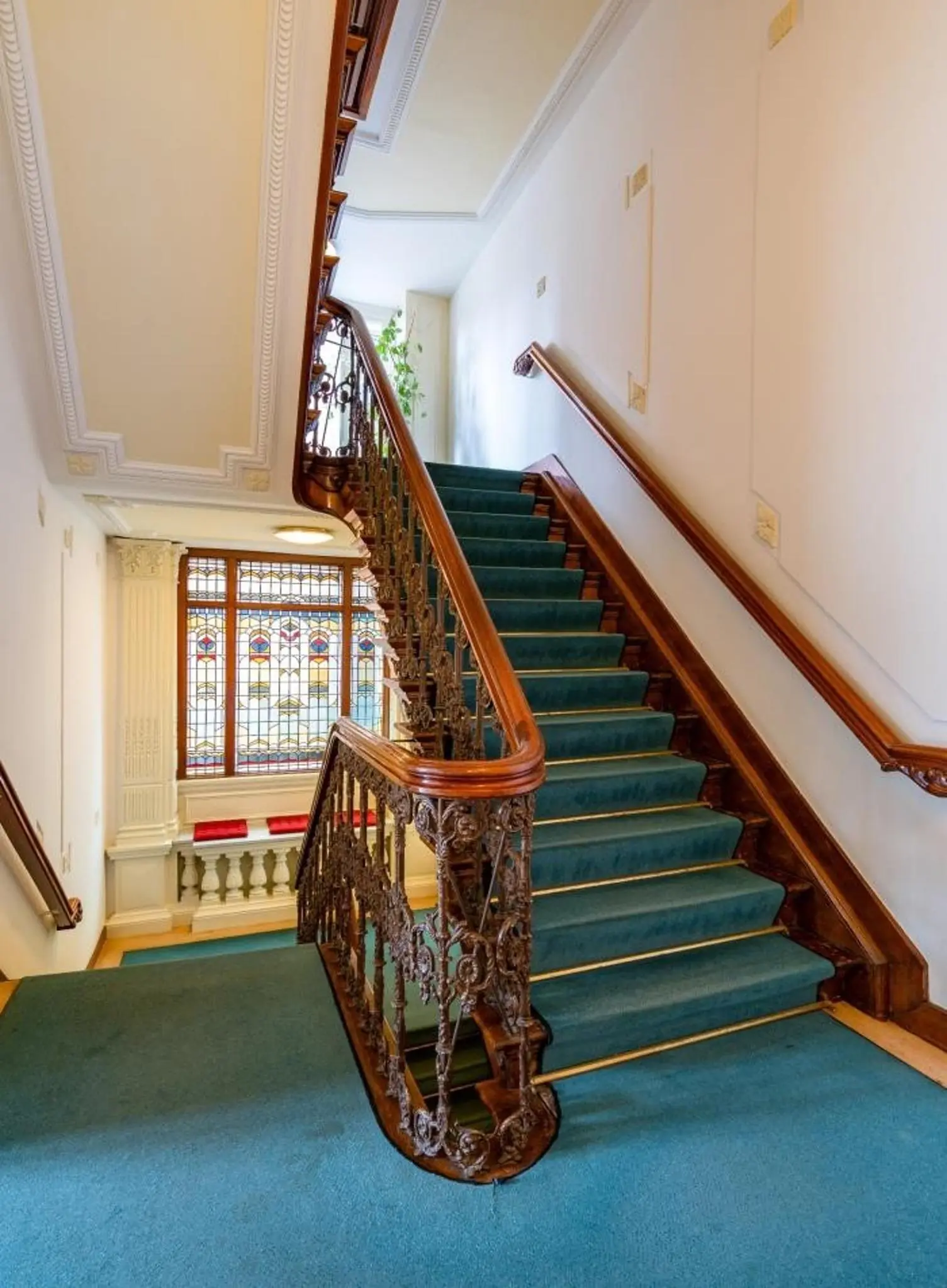 Trap met blauw tapijt en sierlijk houten hekwerk in een klassiek pand aan de Sarphatistraat, met een groot glas-in-loodraam.
