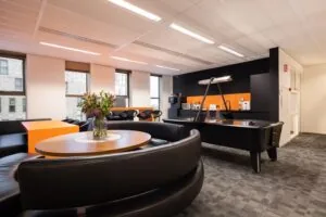 Moderne kantoorlounge met ronde zithoek, bloemen op tafel, airhockeytafel en koffiecorner met oranje accenten.