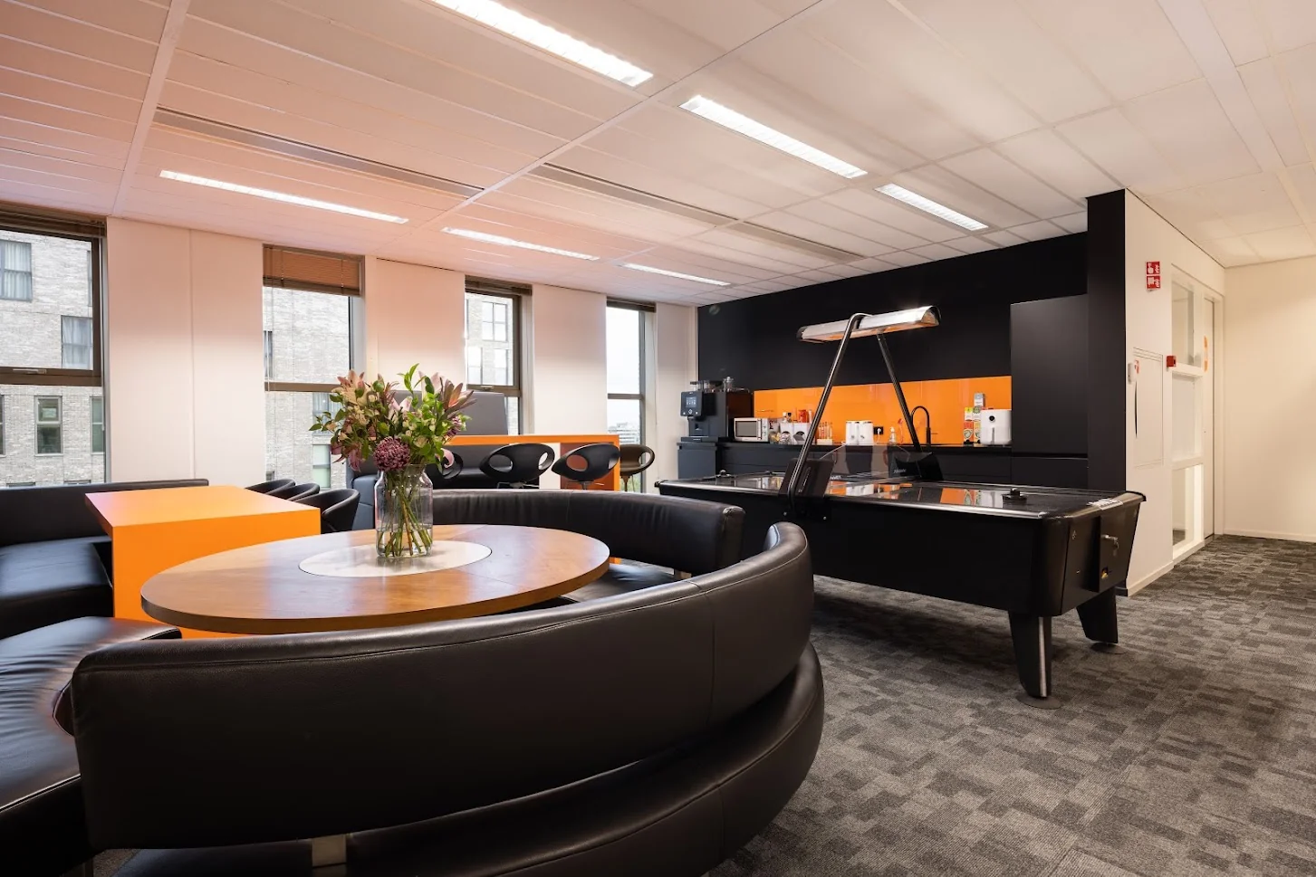Moderne kantoorlounge met ronde zithoek, bloemen op tafel, airhockeytafel en koffiecorner met oranje accenten.
