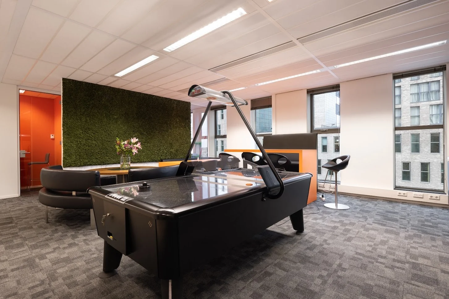 Moderne kantoorruimte met een airhockeytafel, zithoek, groene moswand en grote ramen met uitzicht op omliggende gebouwen.