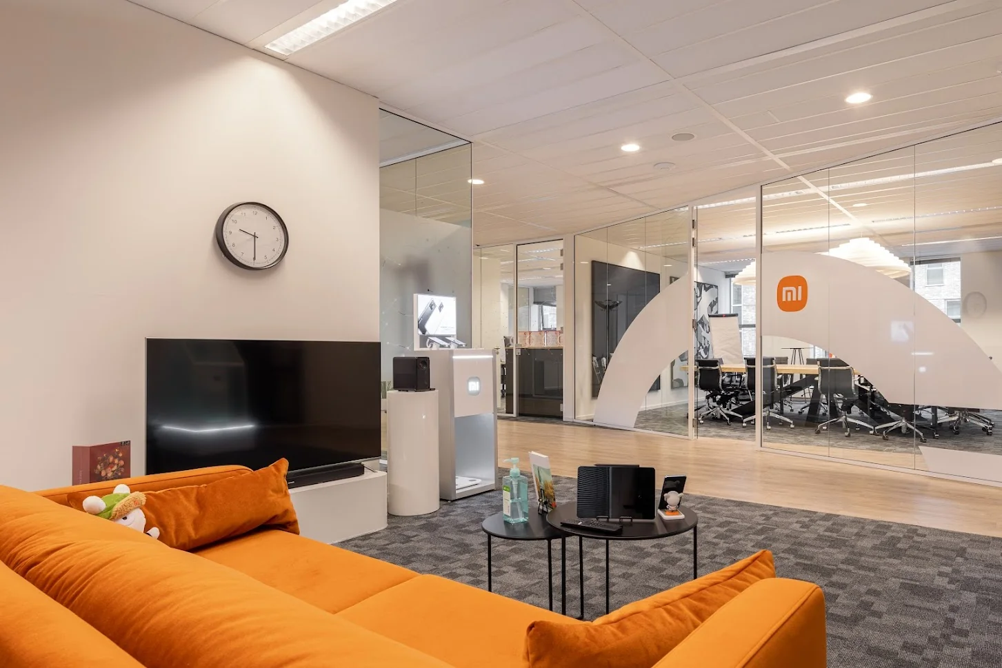 Moderne kantoorruimte van Xiaomi met een oranje bank, flatscreen tv, glazen vergaderruimtes en zichtbaar logo op de glazen wand.
