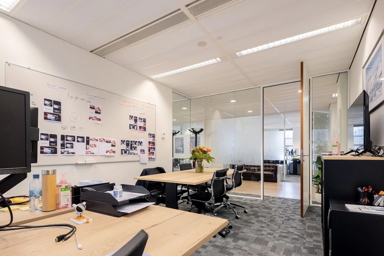 Moderne kantoorruimte met houten bureaus, glazen wanden, een whiteboard vol aantekeningen en een vaas bloemen op de vergadertafel.