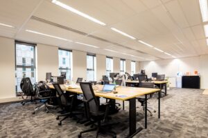 Moderne open kantoorruimte met meerdere bureaus, bureaustoelen, computers en grote ramen die veel daglicht binnenlaten.