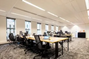 Moderne open kantoorruimte met meerdere bureaus, bureaustoelen, computers en grote ramen die veel daglicht binnenlaten.