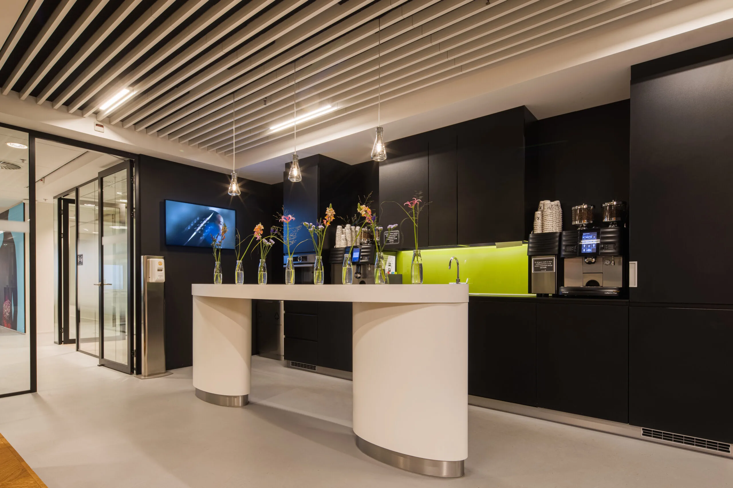 Moderne kantoorpantry met een wit eiland, zwarte kasten, koffieautomaat en decoratieve bloemen in glazen vazen.