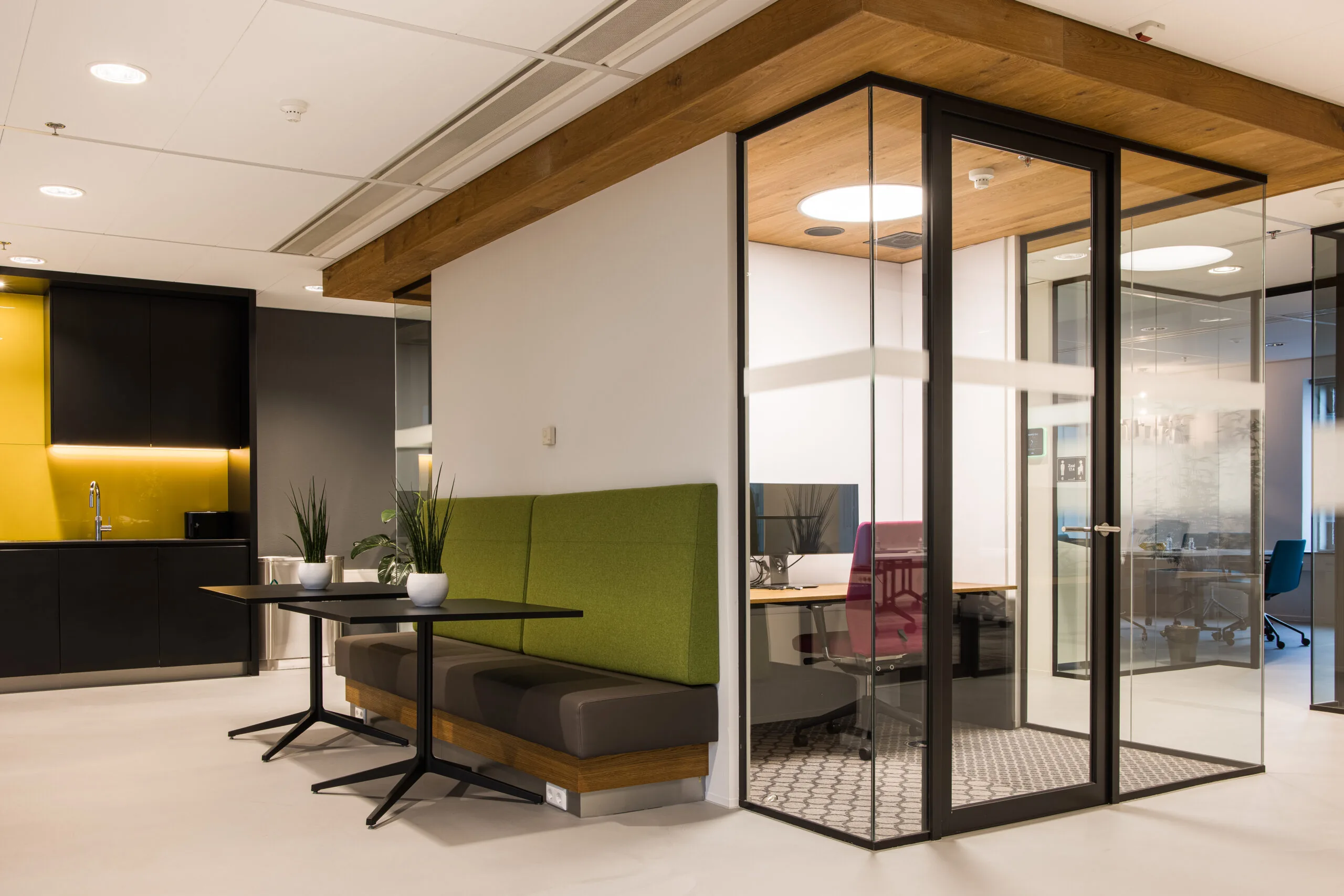 Moderne kantoorruimte met glazen vergaderruimte, zithoek met groene bank en een kitchenette met gele achterwand.