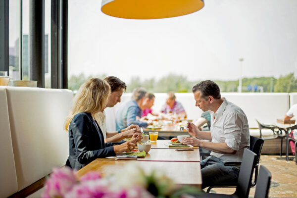 Mensen zitten samen te lunchen aan een lange tafel in een modern bedrijfsrestaurant aan de Orteliuslaan.
