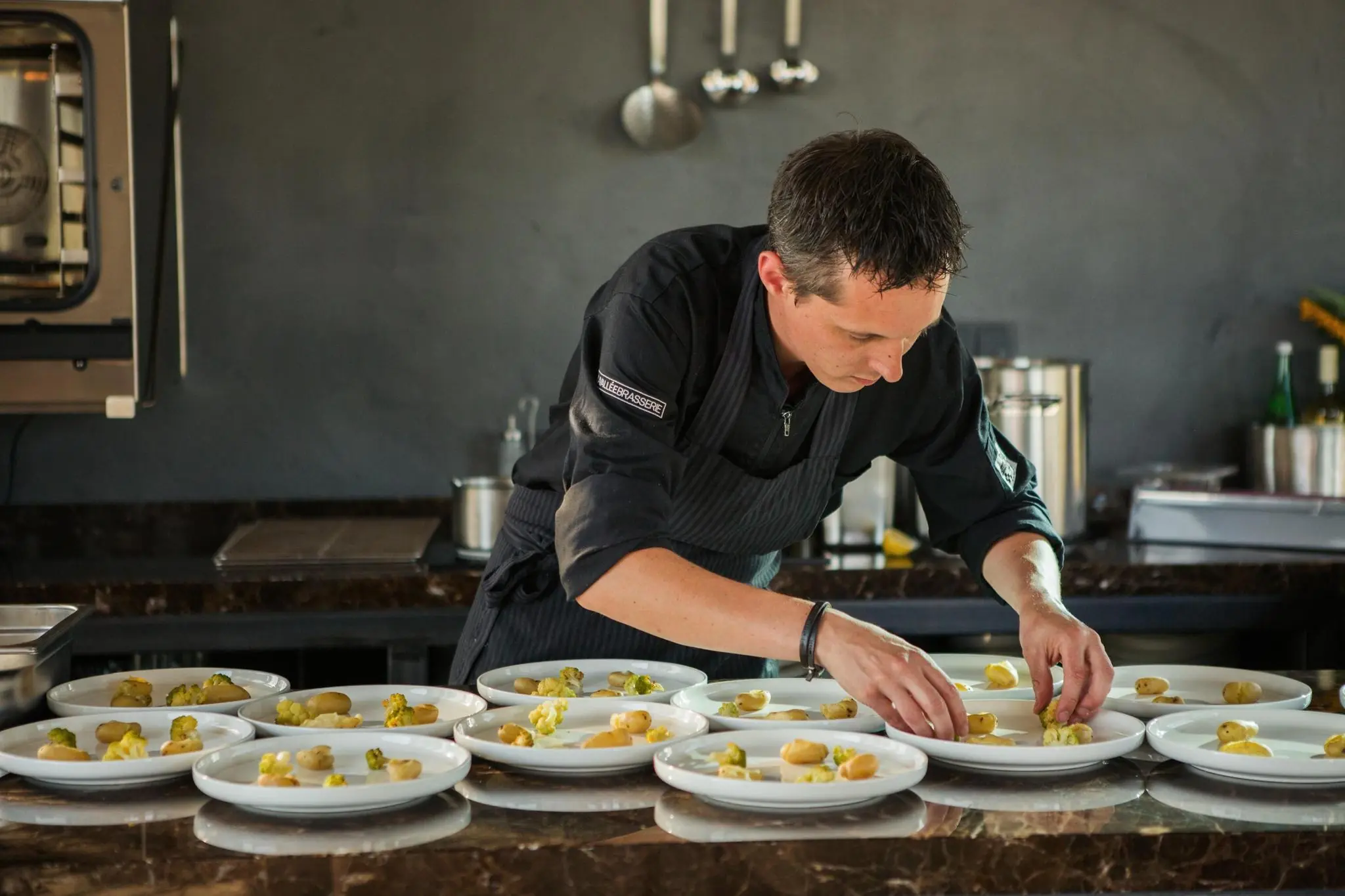 Een chef-kok in zwarte uniform bereidt zorgvuldig meerdere voorgerechten op witte borden in een moderne keuken aan de Orteliuslaan.
