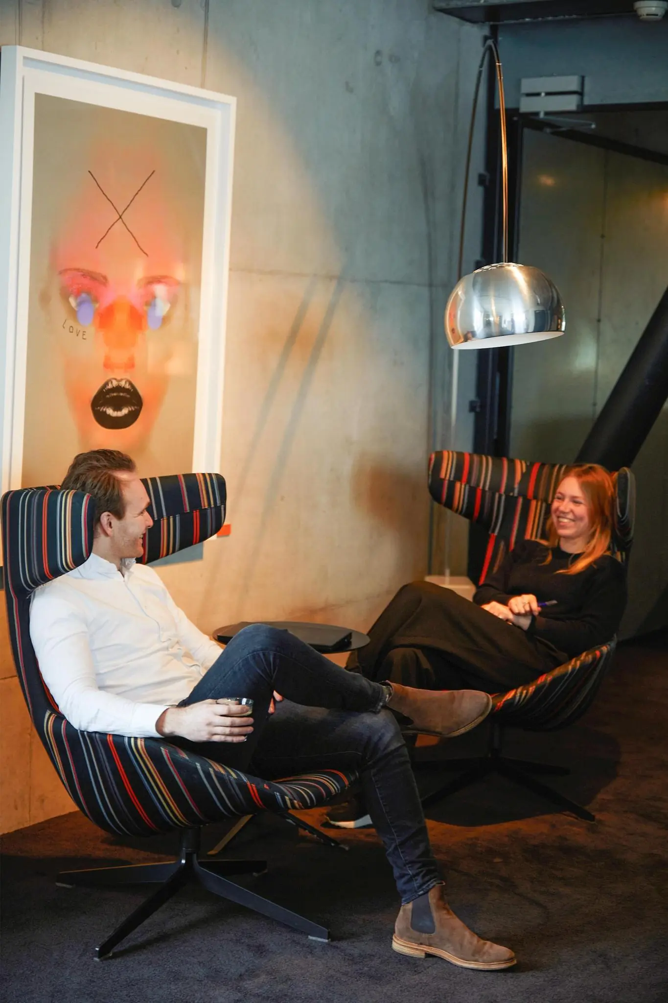 Twee mensen zitten ontspannen in kleurrijke fauteuils onder een moderne lamp bij Orteliuslaan, met een kunstwerk op de achtergrond.