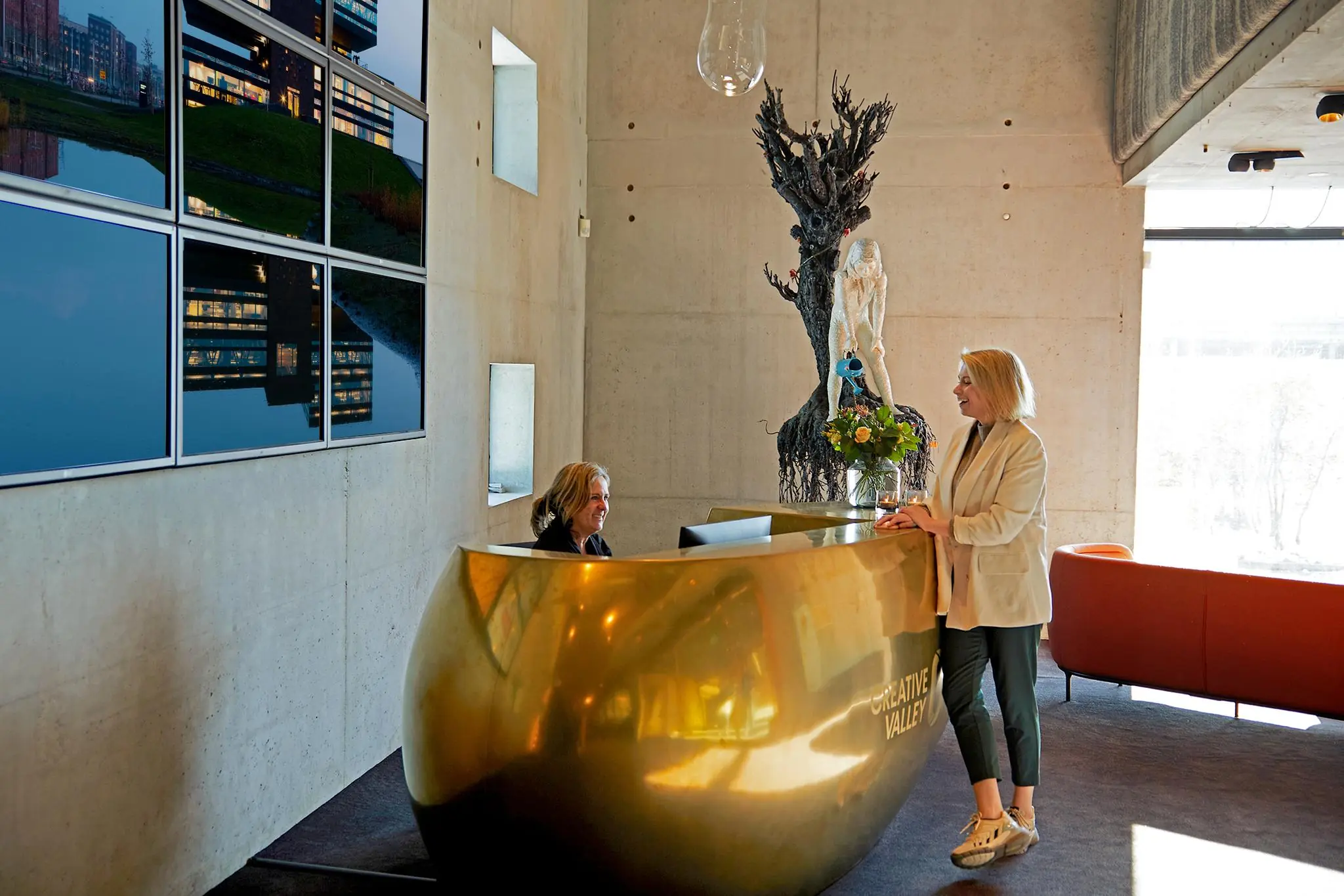 Vrouw bij de receptie van Creative Valley aan de Orteliuslaan, met gouden balie, kunstwerk en moderne inrichting.