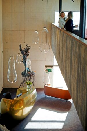 Moderne ontvangsthal aan de Orteliuslaan met een goudkleurige balie, hangende glazen lampen en twee vrouwen die praten op de bovenverdieping.