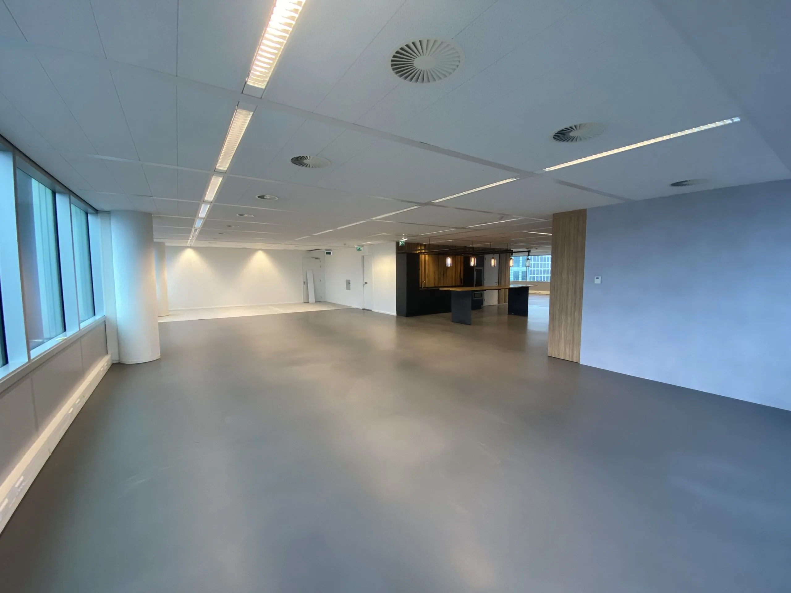 Moderne, lege kantoorruimte met grijsvloer, grote ramen en een open keuken in de hoek.