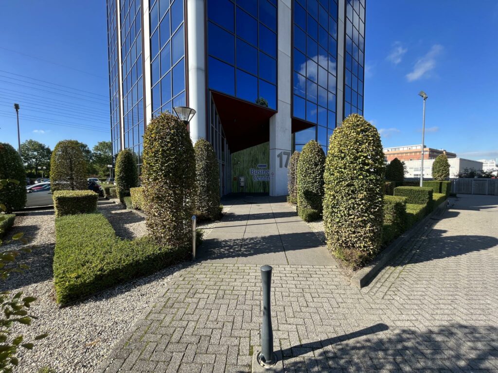 Ingang van een modern kantoorgebouw aan de Savannahweg 17 met strak gesnoeide hagen en glazen gevel.