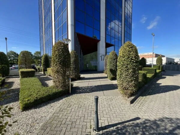 Ingang van een modern kantoorgebouw aan de Savannahweg 17 met strak gesnoeide hagen en glazen gevel.