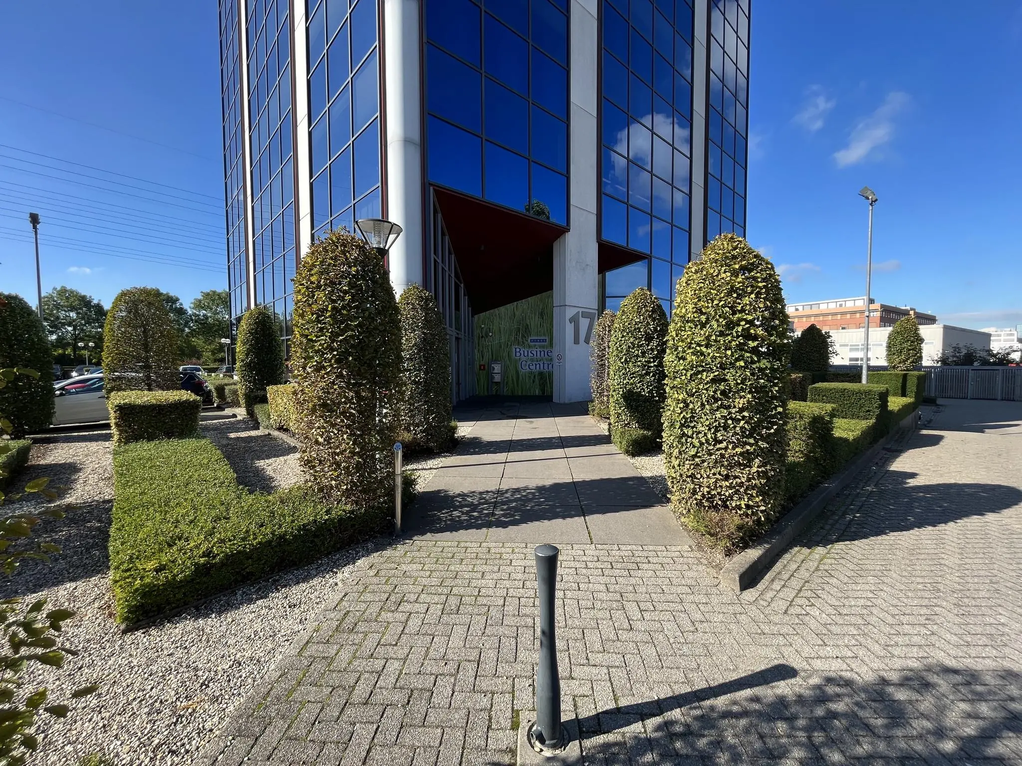 Ingang van een modern kantoorgebouw aan de Savannahweg 17 met strak gesnoeide hagen en glazen gevel.