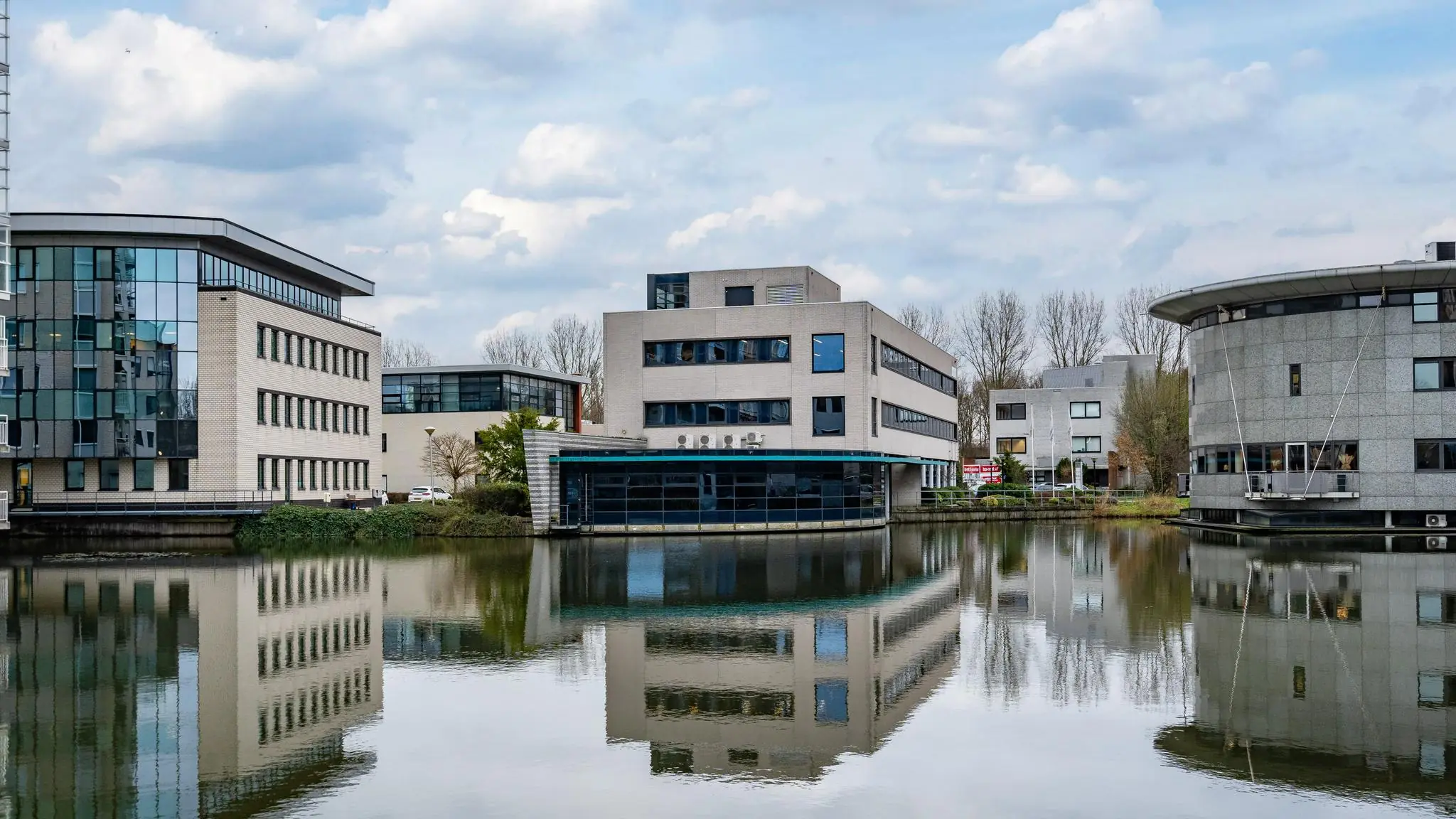 Kantoorpanden aan de Barbizonlaan weerspiegelen in het water onder een deels bewolkte hemel.