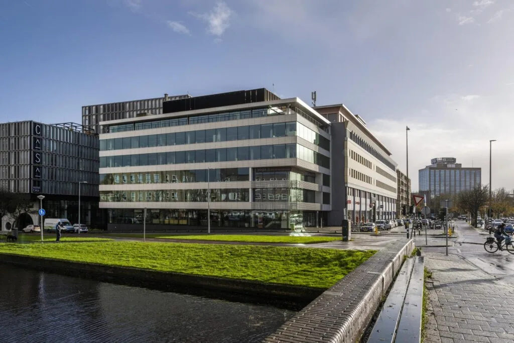 Straatbeeld van de Wibautstraat in Amsterdam met moderne kantoorgebouwen, waaronder Hotel Casa, en een groenstrook langs het water op de voorgrond.
