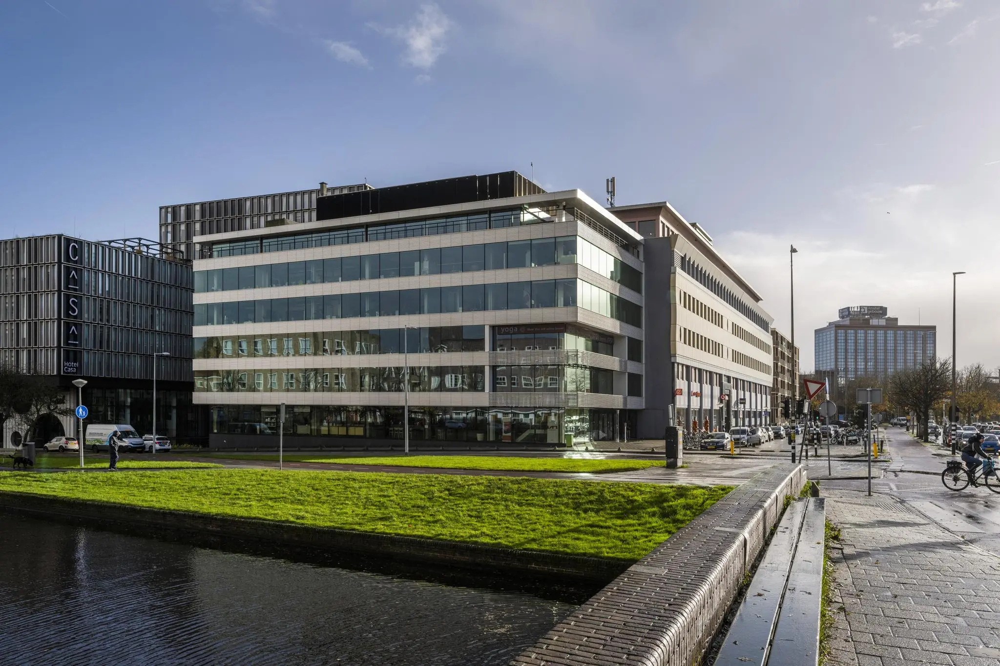 Straatbeeld van de Wibautstraat in Amsterdam met moderne kantoorgebouwen, waaronder Hotel Casa, en een groenstrook langs het water op de voorgrond.