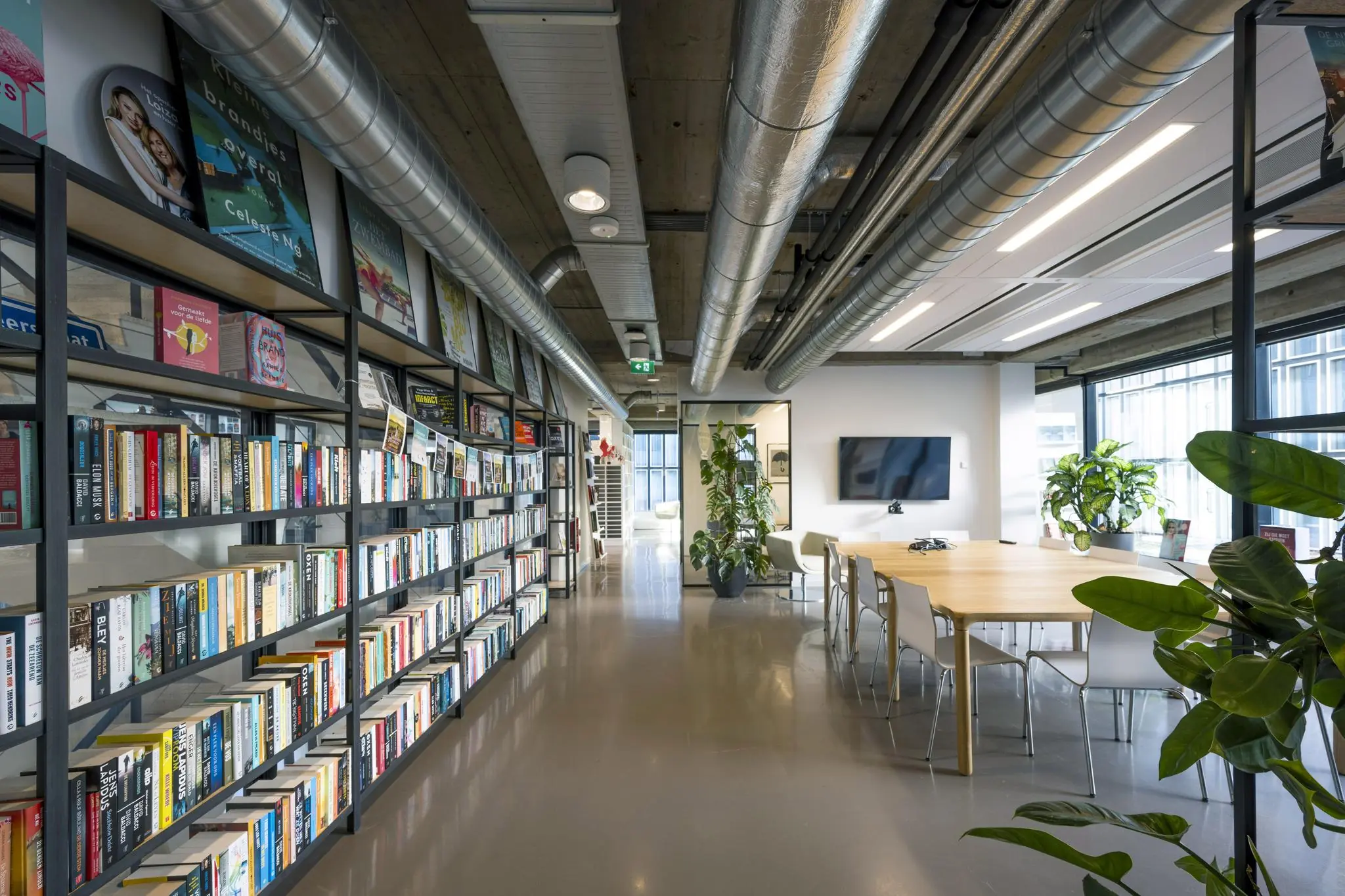 Moderne kantoorruimte aan de Wibautstraat met een lange boekenkast, vergadertafel, planten en industriële plafonddetails.