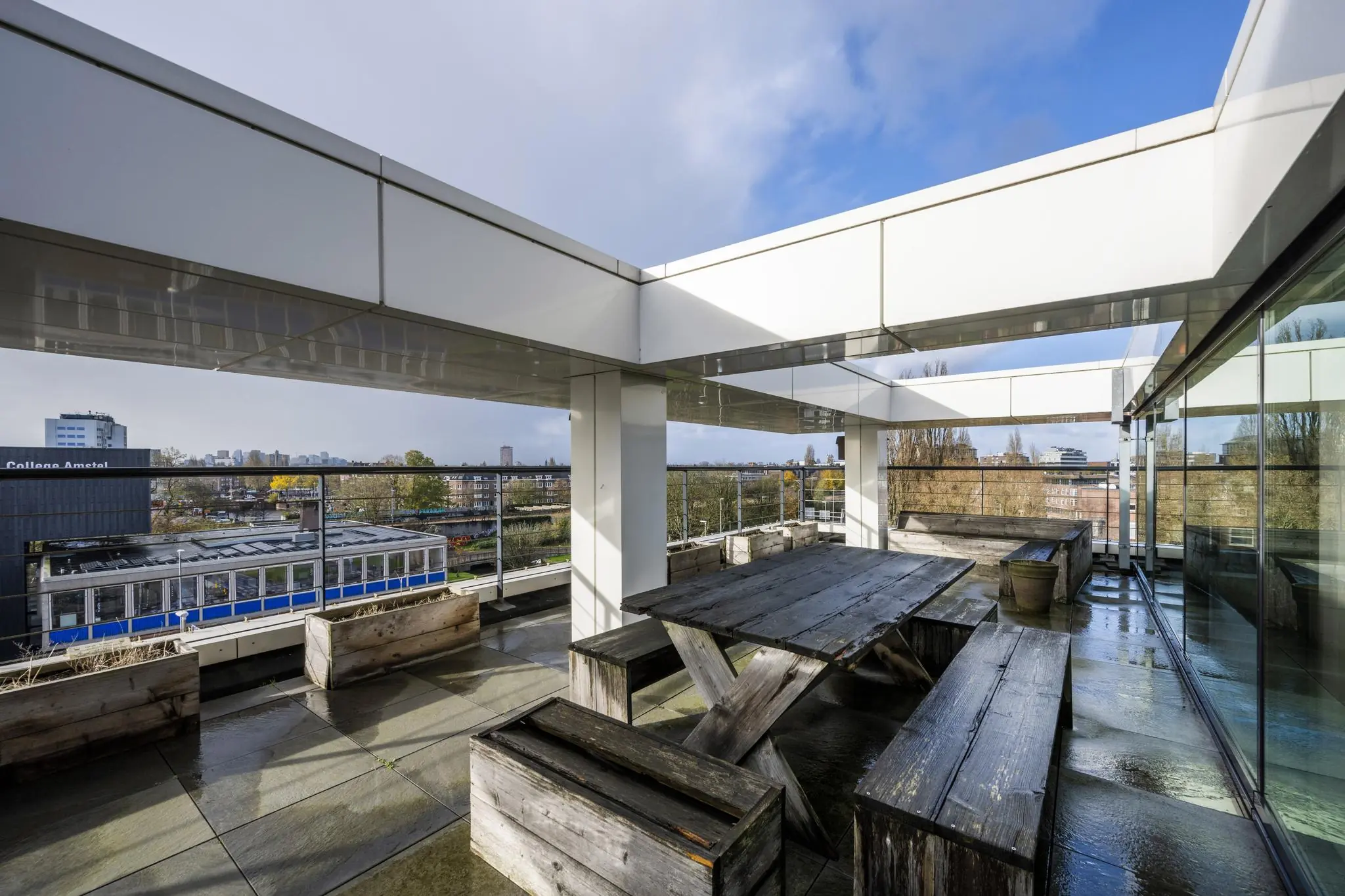 Dakterras met houten picknicktafels op een modern gebouw aan de Wibautstraat in Amsterdam, met uitzicht op de stad en een passerende tram.