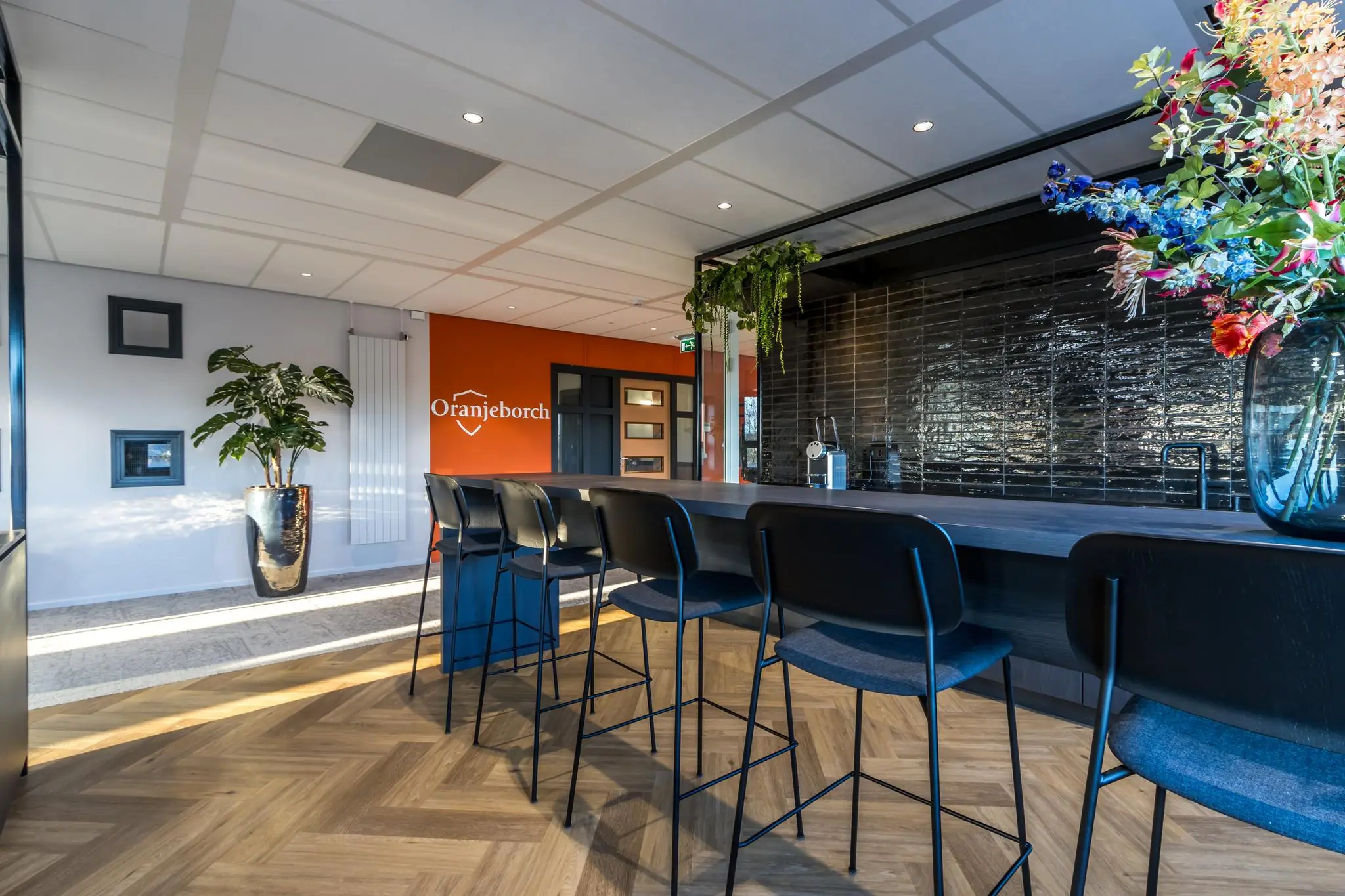 Moderne ontvangstruimte van Oranjeborch met hoge zitplaatsen aan een bar, opvallende bloemen en een oranje accentmuur.