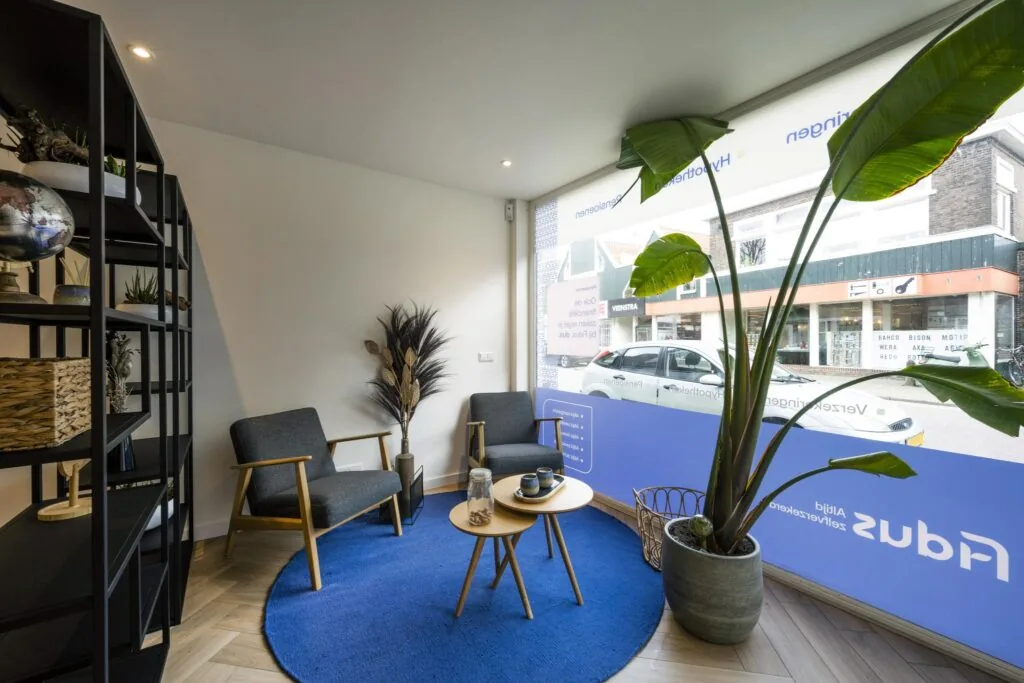 Wachtruimte met twee stoelen, een plant en uitzicht op de Raadhuisstraat in een modern ingerichte praktijkruimte.