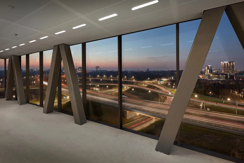 Uitzicht vanuit een modern kantoorgebouw aan de Joan Muyskenweg op een snelwegknooppunt bij zonsondergang.
