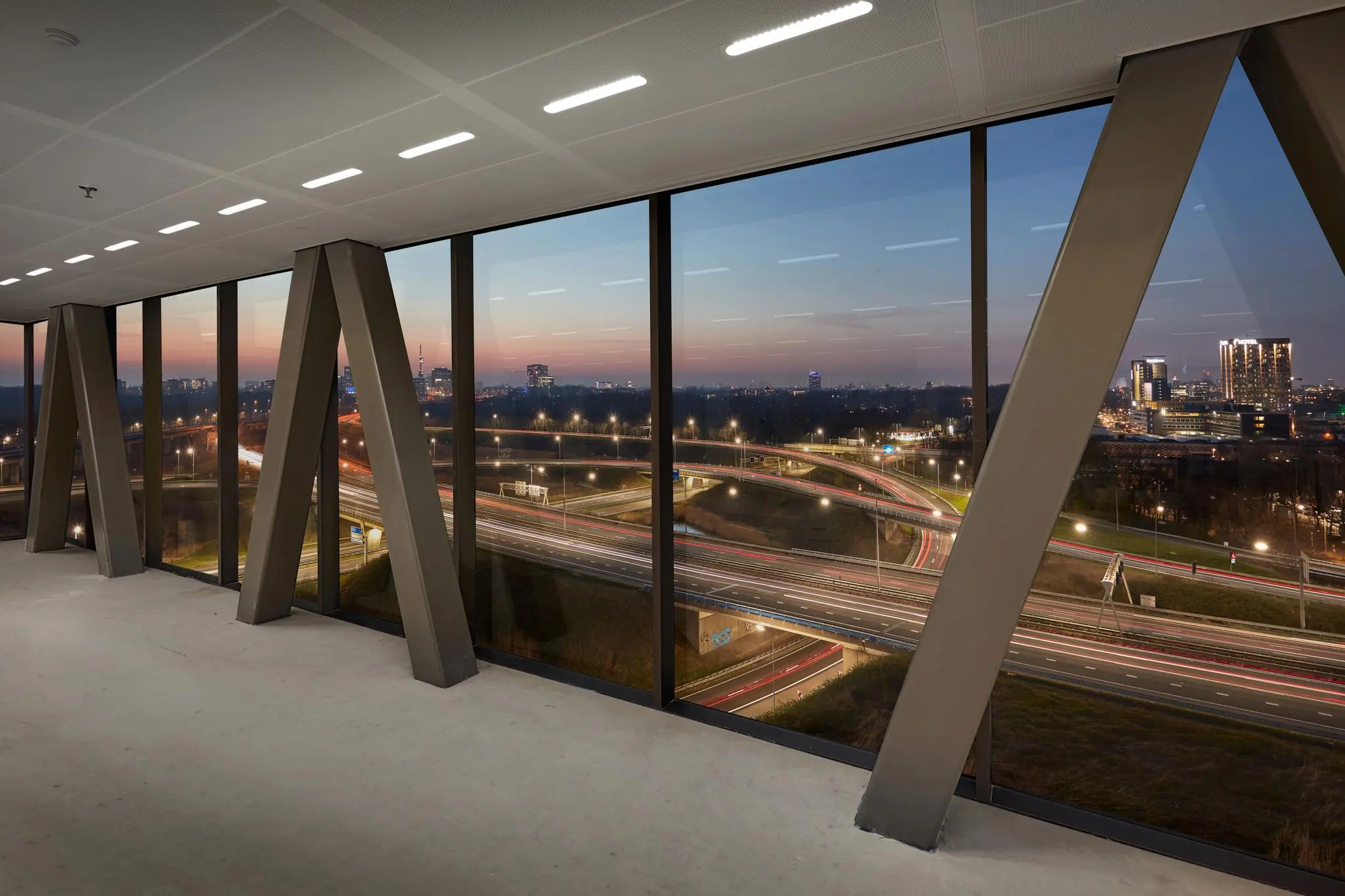 Uitzicht vanuit een modern kantoorgebouw aan de Joan Muyskenweg op een snelwegknooppunt bij zonsondergang.