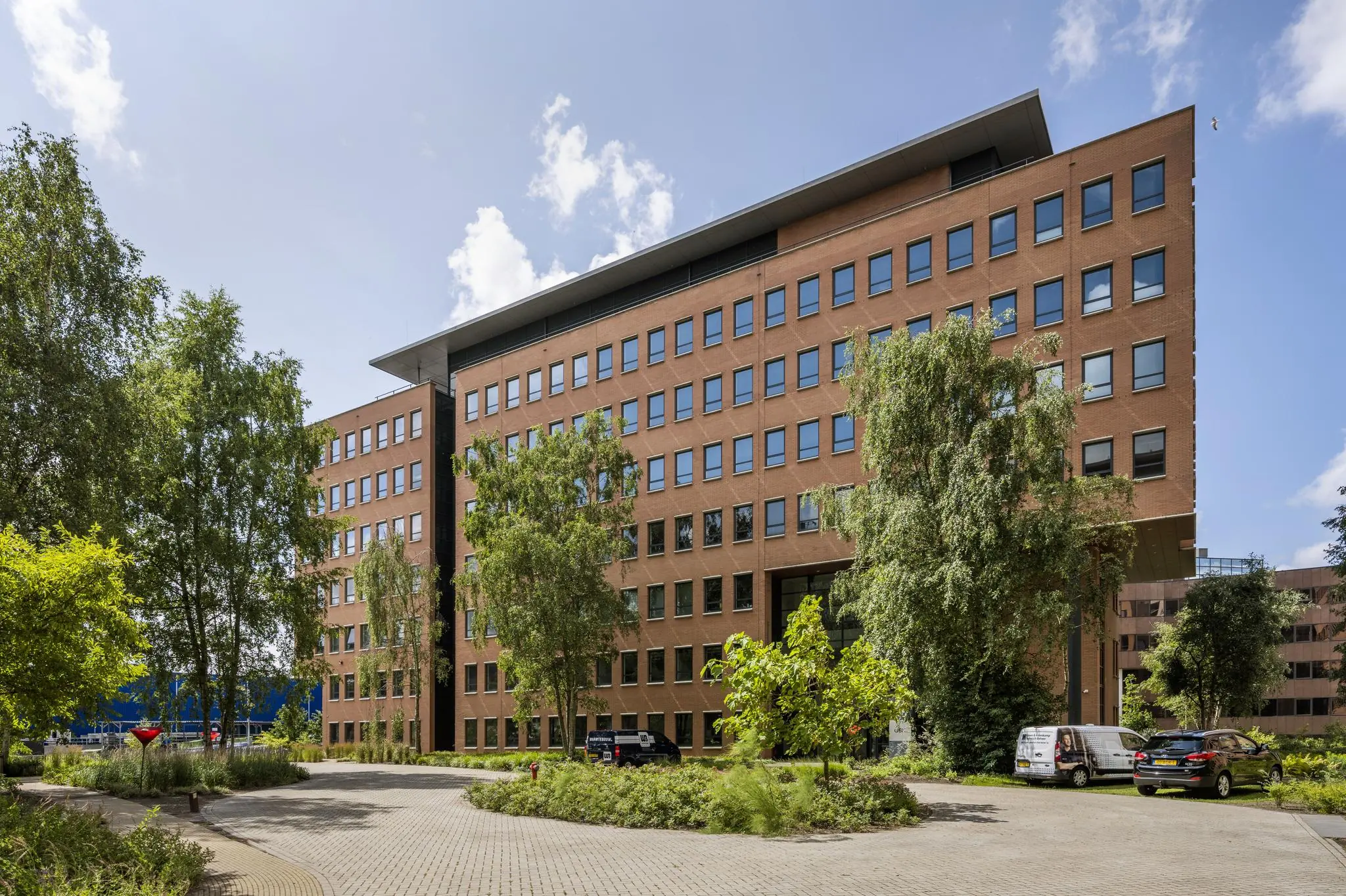 Kantoorgebouw aan de Hullenbergweg, omringd door groen en geparkeerde auto's op een zonnige dag.