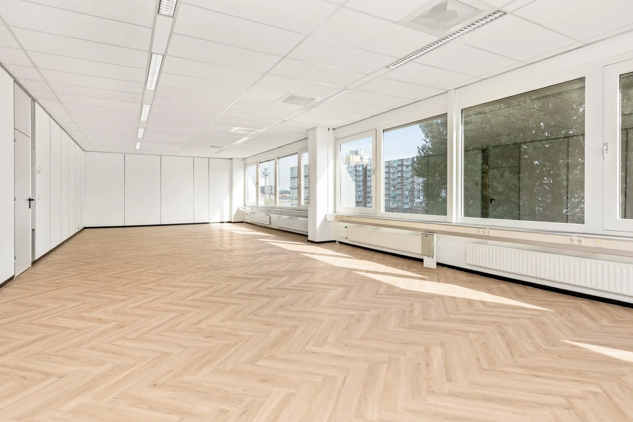 Lichte, lege kantoorruimte aan de Sluisjesdijk met visgraatvloer en grote ramen met uitzicht op hoogbouw.