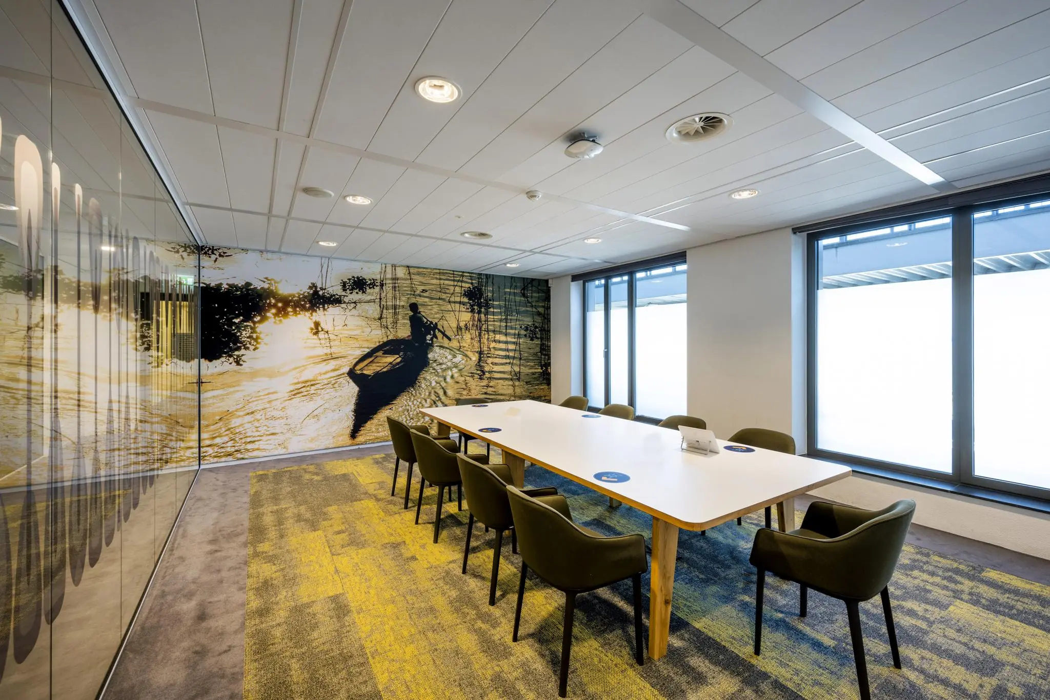 Vergaderruimte aan de Piet Heinkade met een grote tafel, groene stoelen en een fotowand met een roeiboot op het water bij zonsondergang.