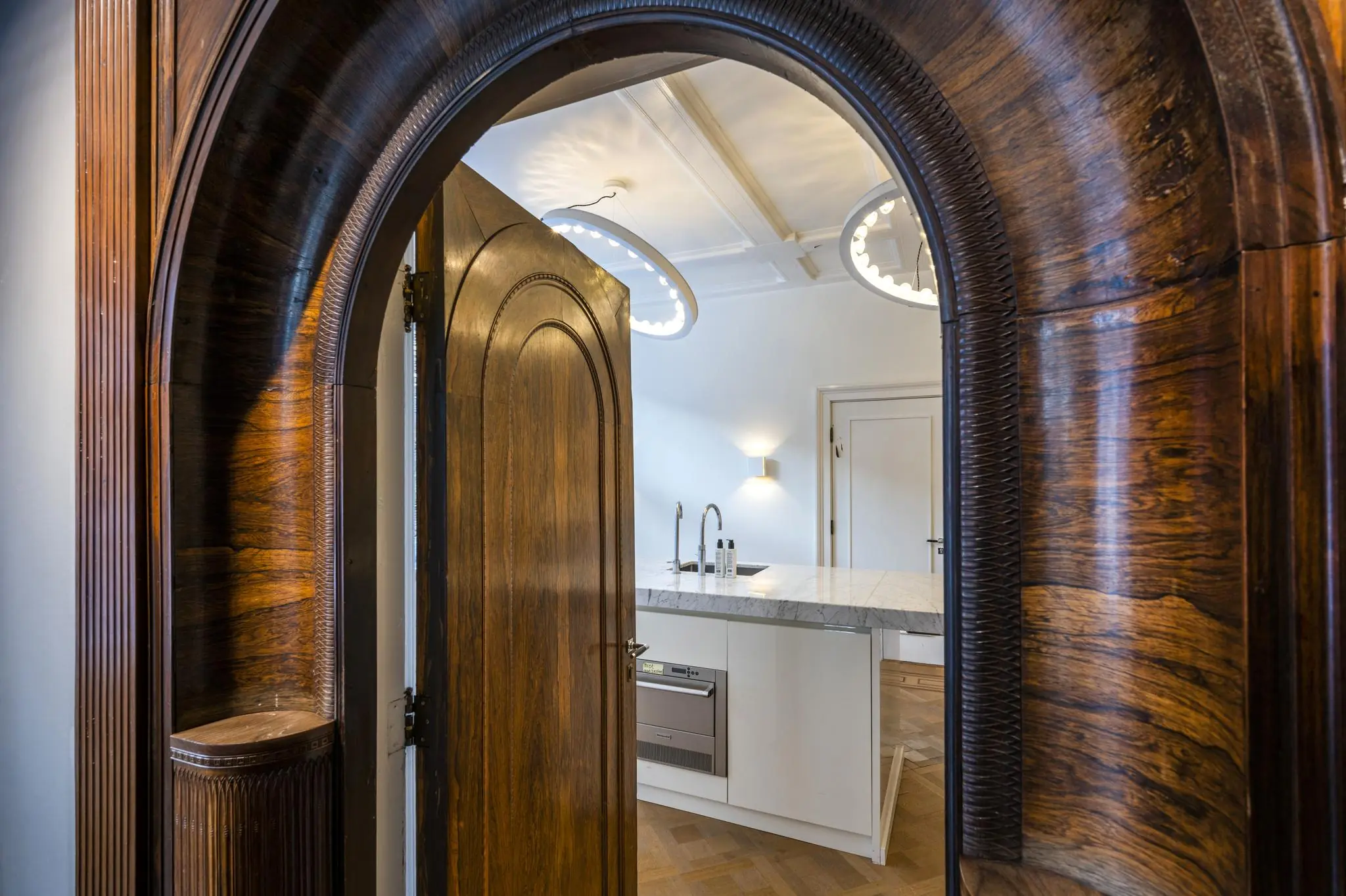 Doorkijk door een klassieke houten boogdeur naar een moderne keuken met marmeren aanrechtblad in de Honthorststraat.