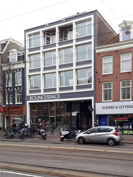 Een moderne vier verdiepingen tellend gebouw met de naam "BounceSpace" op de begane grond, omringd door fietsen en geparkeerde auto's.