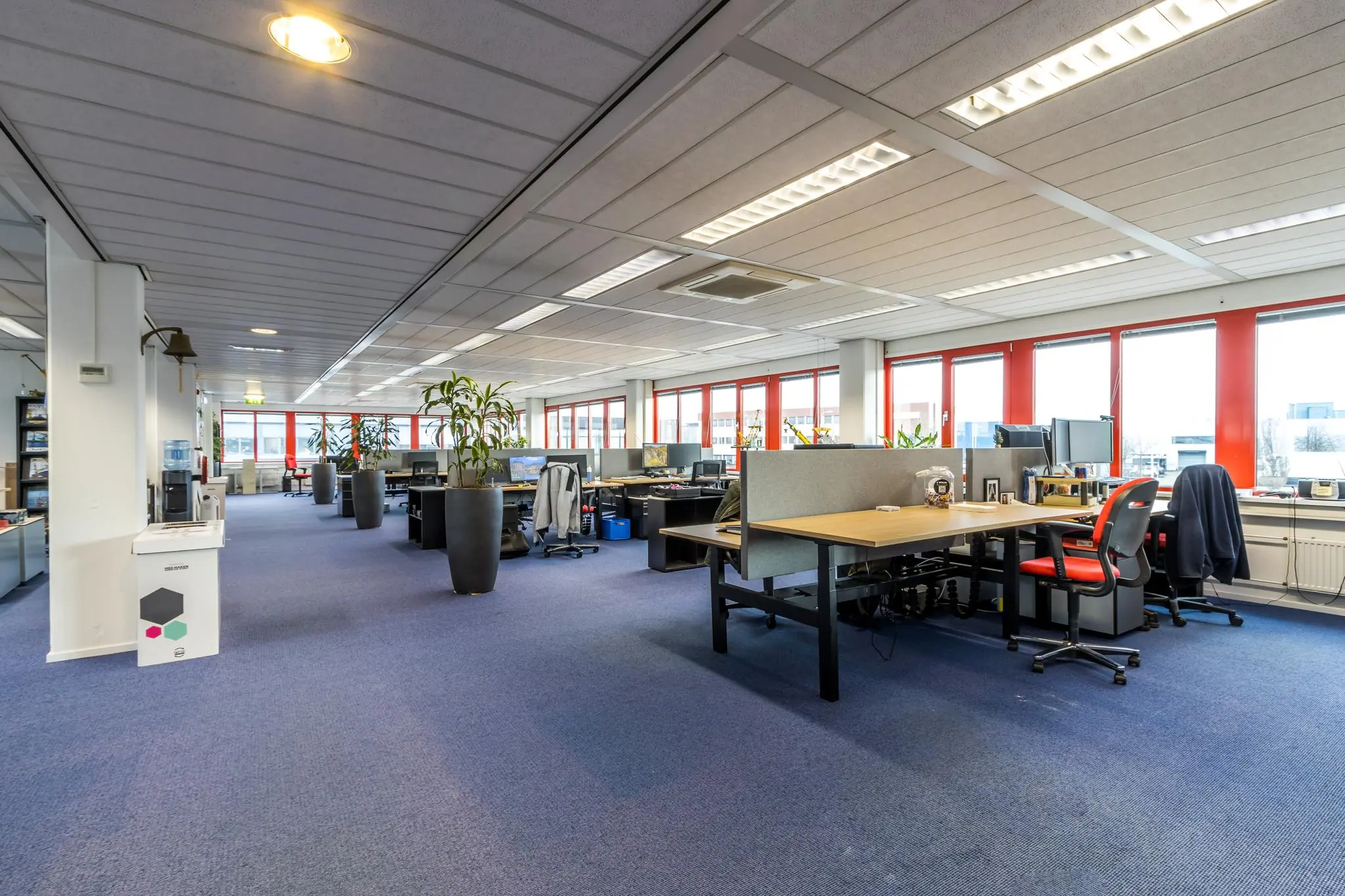 Ruim en licht kantoorpand aan de Savannahweg met open werkplekken, rode raamkozijnen en blauwe vloerbedekking.