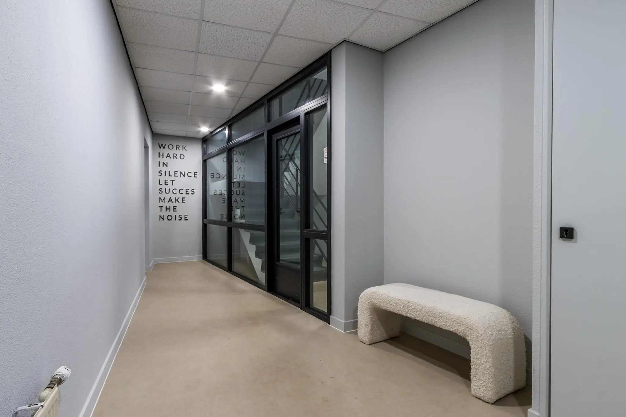 Moderne gang met glazen wand, witte bank en motiverende tekst op de muur in een gebouw aan de Koningsweg.