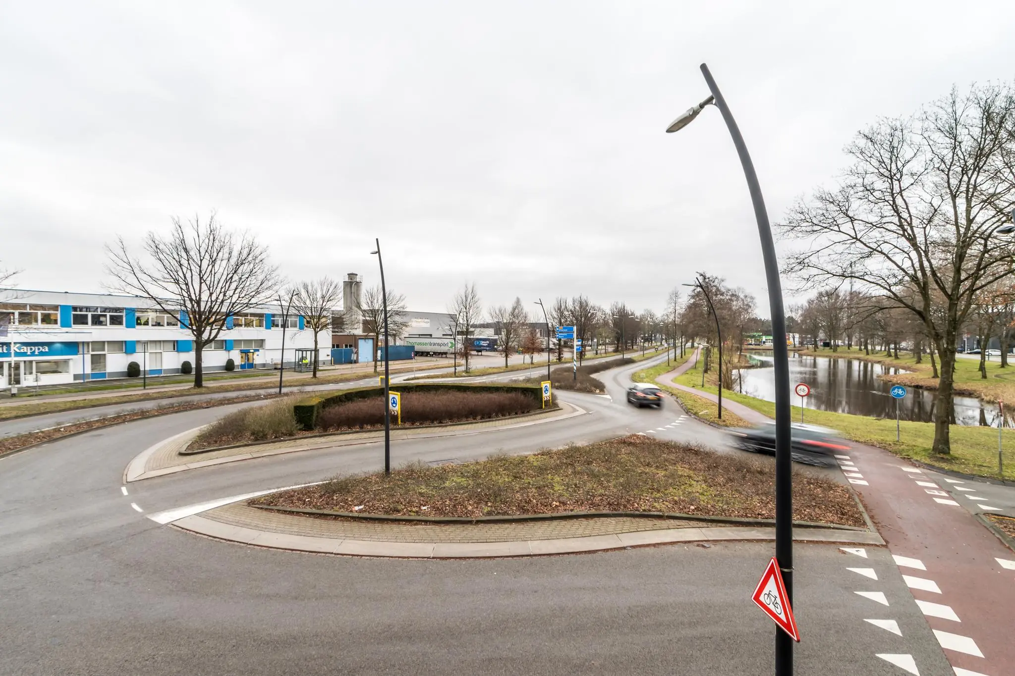 Rotonde aan de Koningsweg met voorbijrijdende auto's, fietspad en bedrijven zoals Smurfit Kappa op de achtergrond.