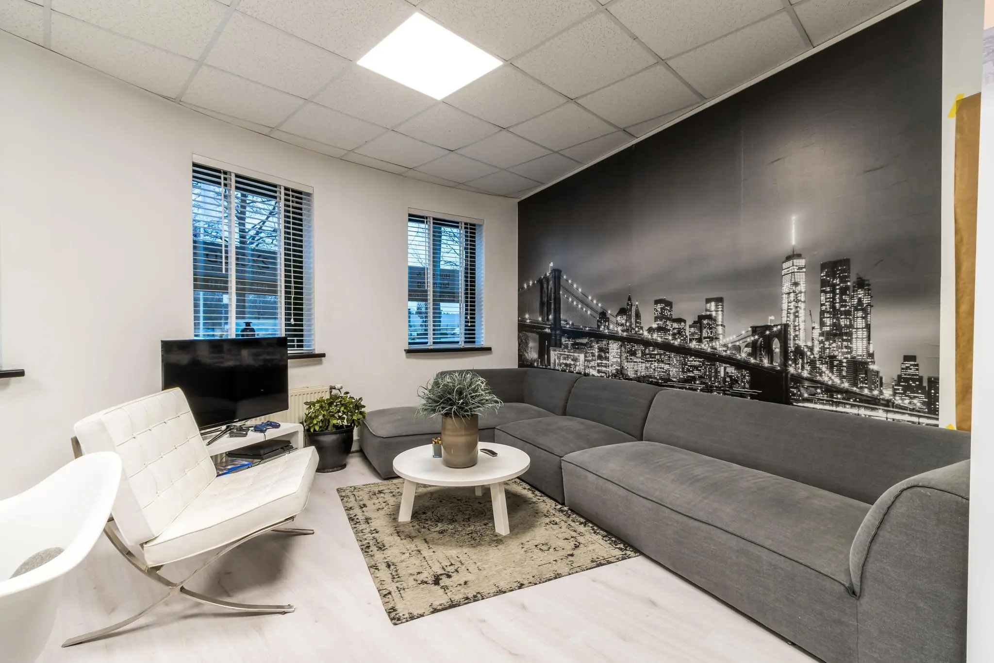 Moderne zithoek met grijze hoekbank, witte stoelen en een muurfoto van de skyline van New York aan de Koningsweg.