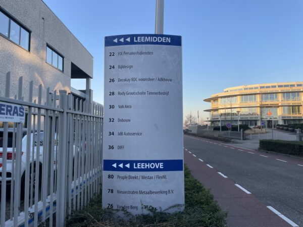 Wegwijzer met bedrijfsnamen en huisnummers op bedrijventerrein Leemidden en Leehove in een stedelijke omgeving.