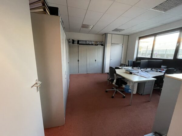 Kantoorruimte met bureaus, kantoormeubilair, archiefkasten en computers aan een roodtapijtvloer.